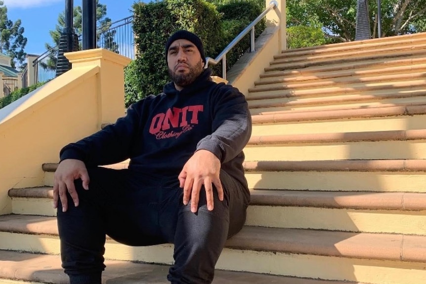 Man in black beanie, black hoodie, pants and red sneakers sits on orange staircase outdoors. Green shrub fringes the stairtops