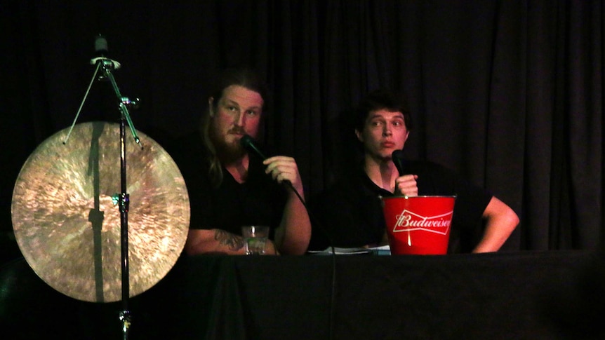 Two men sit on stage with a gong next to them.