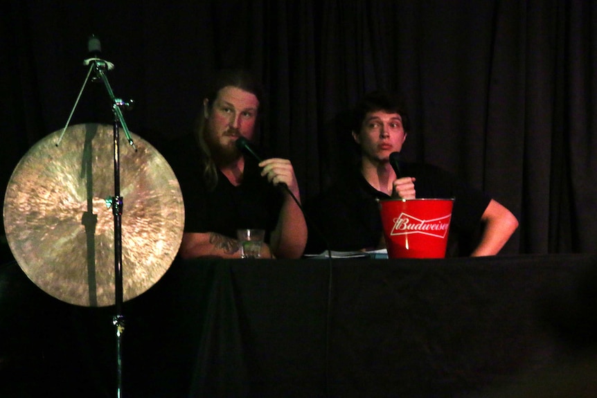 Two men sit on stage with a gong next to them.