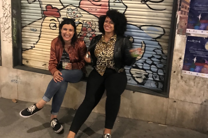 Two women are seen sitting on a ledge against a wall with graffiti. They both look to the camera and smile, it's night.