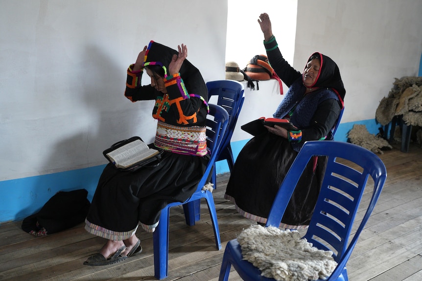 Two women sit on plastic chairs with bibles on their laps, praying with their arms outstretched upwards. 