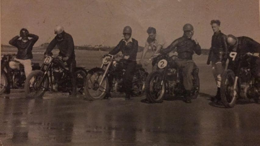 An old photo of five men on old school motorbikes parked on a beach.