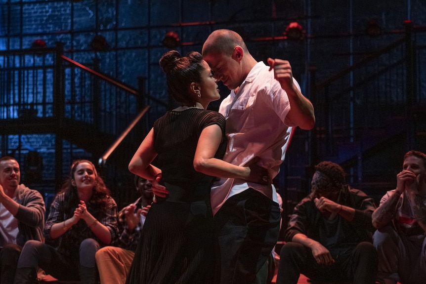 On stage, a Latino woman stands close to a muscular man with a buzz cut. She is holding him at his sides.