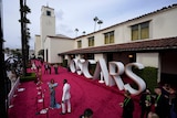 A woman in a blue gown and a man in a white suit stand on an empty pink carpet with an Oscars sign behind them.