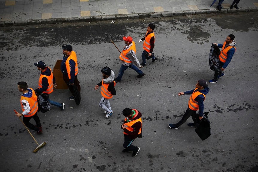 A group of young people wearing orange vests walk down a street and pick up bottles and other rubbish following large protests.