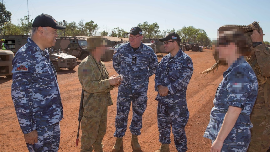 Three male politicians stand in blue camouflage outfits and speak with Defence officials