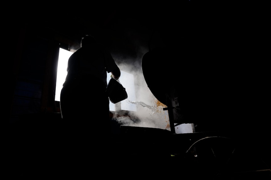 A silhouette of a man throws water from a bucket over steaming apparatus in a dark shed