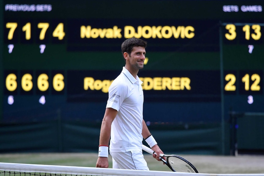 A male tennis player holding a racquet in his left hand smiles as he walks in front of a scoreboard.