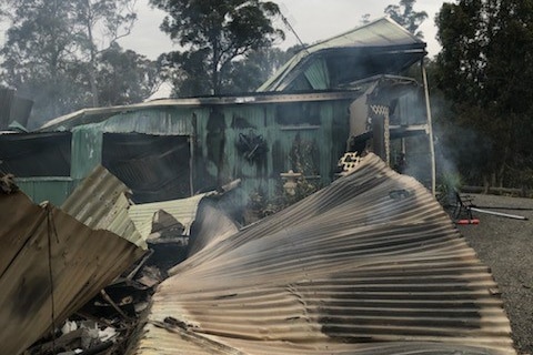 A fence fallen to the right and the charred, smoky remains on a house.