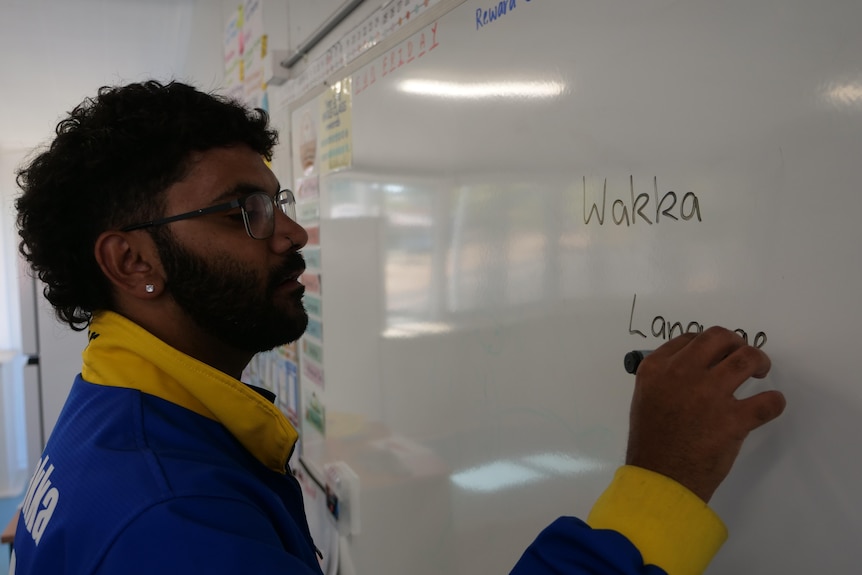 An Indigenous man, beard, glasses, small ear stud, writes Wakka Language' on a whiteboard, wears yellow and blue jersey.