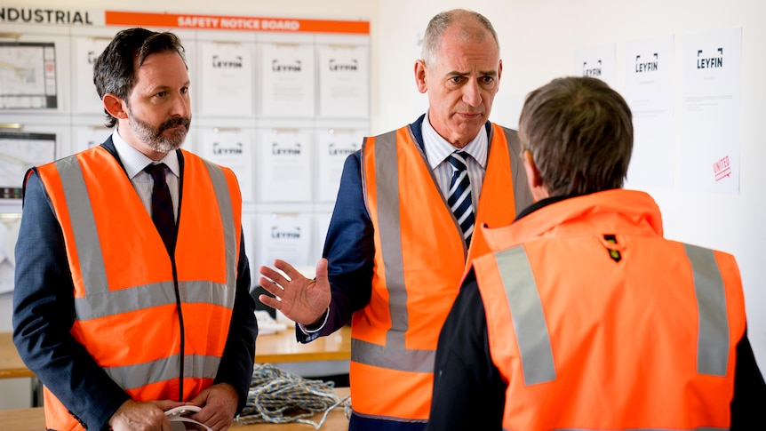 Three men wearing hi-vis orange vests in an office.