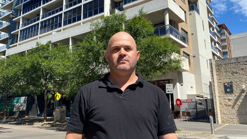 Man dressed in dark polo top stands outside an apartment building.