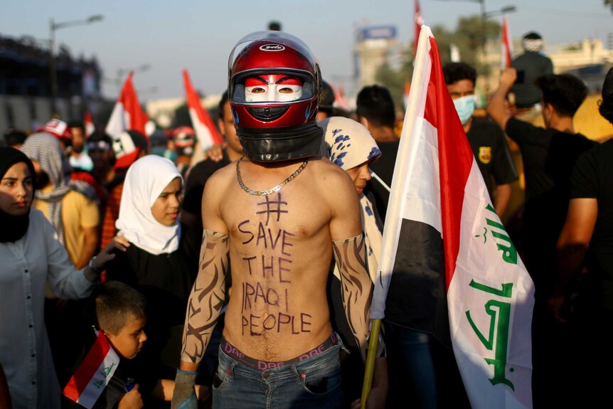 A protester in a red helmet and underneath that a white and red mask, with no shirt has #savetheiraqpeople written on his chest.
