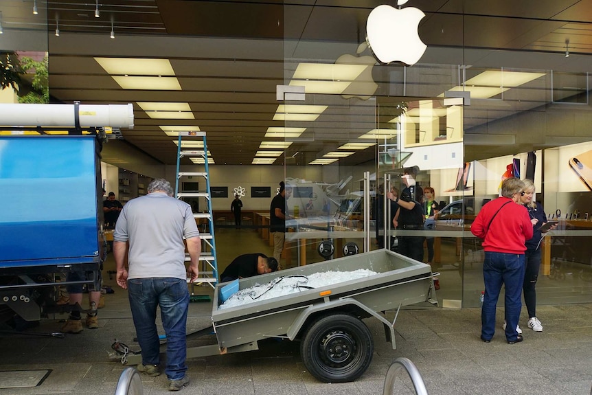 A wide shot showing people and a trailer filled with broken glass outside the Apple Perth City store with a big window missing.