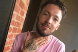 Mitchell Joseph Brindley smiles for a photo sitting at a table in a pink shirt holding a cocktail with one hand on his neck.