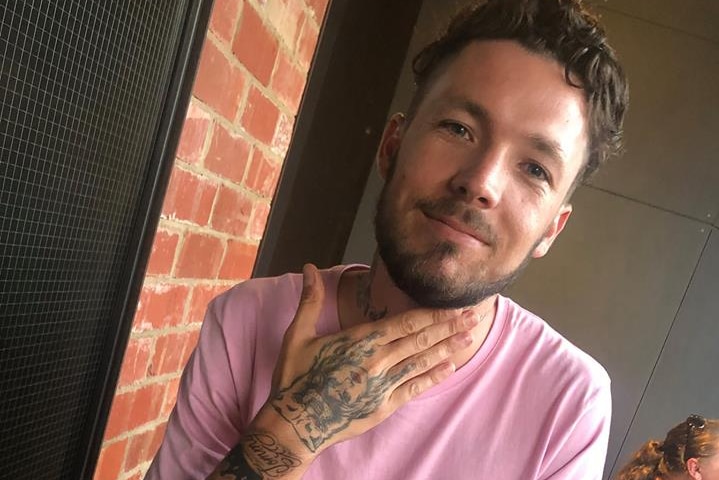 Mitchell Joseph Brindley smiles for a photo sitting at a table in a pink shirt holding a cocktail with one hand on his neck.