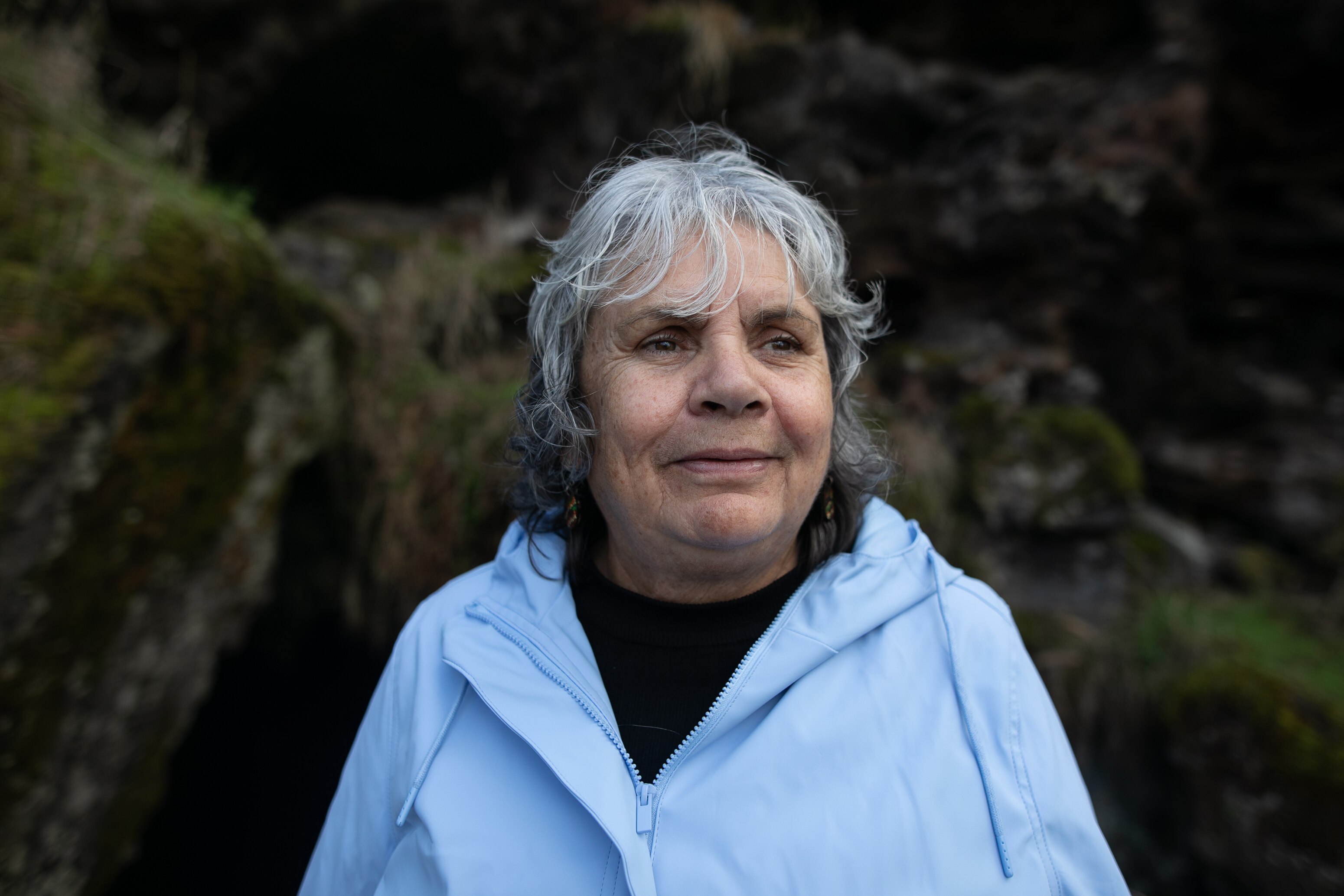 A woman with grey shoulder length hair in pale light blue windbreaker stands look into distance, behind her mossy rocky wall