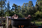 Two young men relax in a hot tub over a fire, the bush all around.