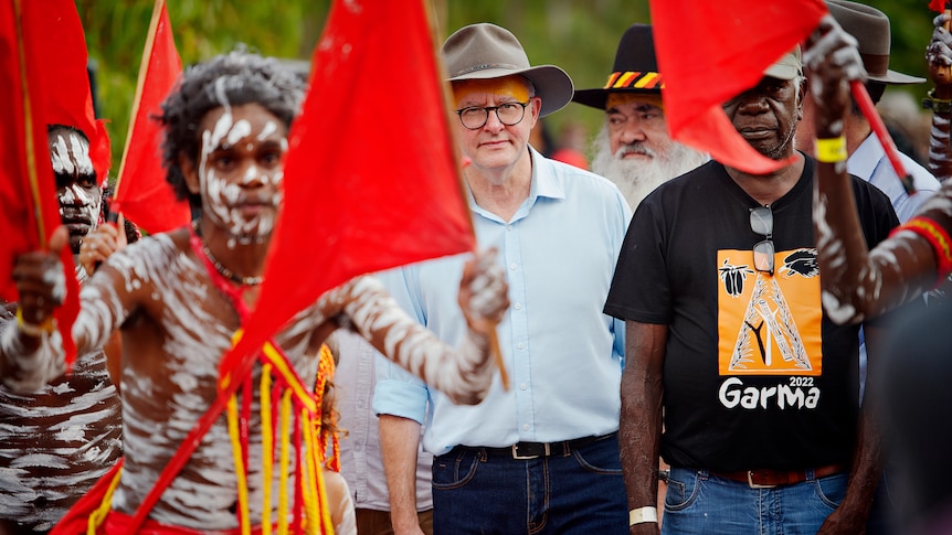 White man with hat walks in the middle of a group of Indigenous people in traditional dress carrying red flags
