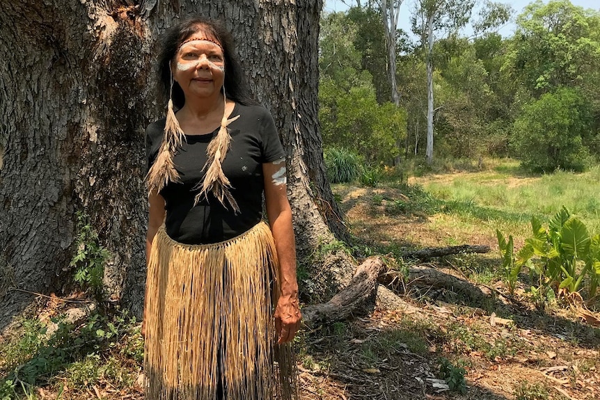 An Aboriginal woman with ochre painted on her cheeks wearing grass skirt stands in front of big tree.