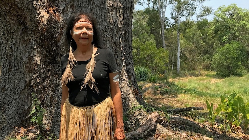 An Aboriginal woman with ochre painted on her cheeks wearing grass skirt stands in front of big tree.