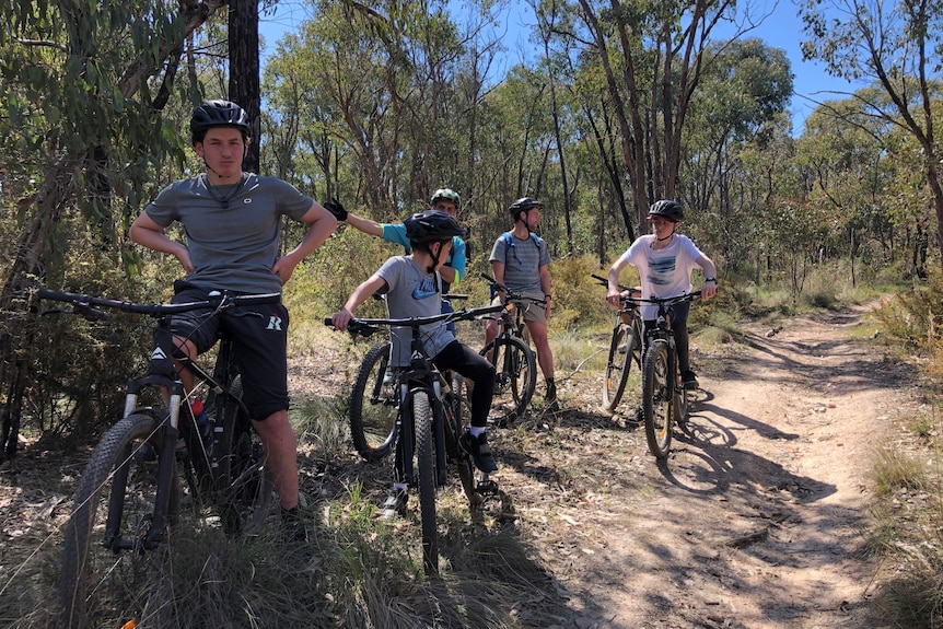 Three teenage boys and two male adults on mountain bikes paused mid-ride next to a bush track.