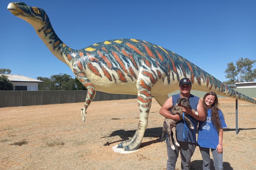 A man and woman standing in front of a dinosaur replica, holding their pet dog.