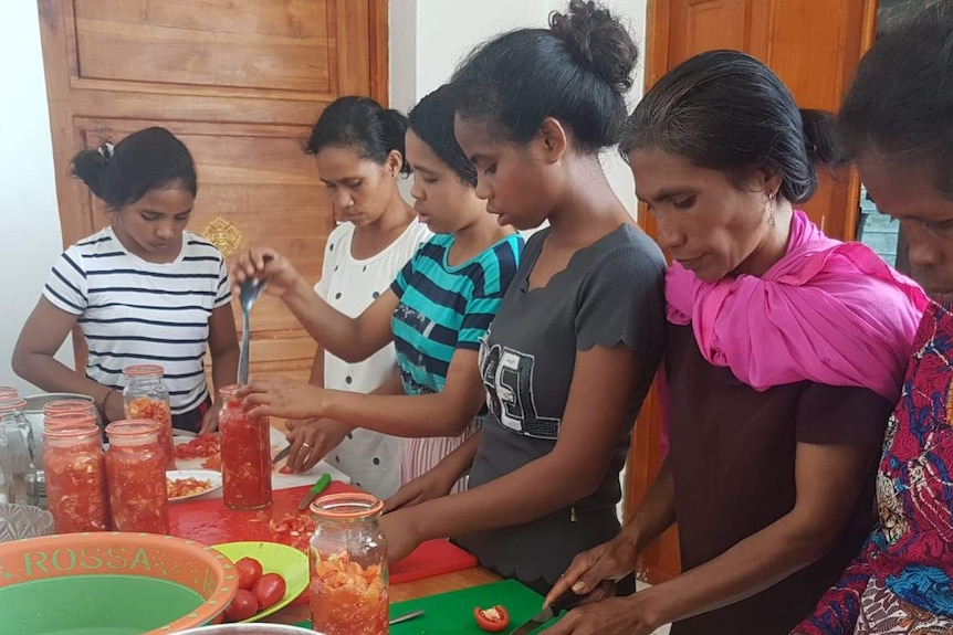 A group of women chop tomatoes and put them in jars around bench covered in jars, bowls, and chopping boards.