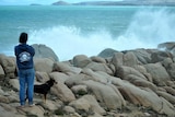 A person in a hoodie, holding a dog on a leash, looks out to sea as foam explodes up over a breakwall.