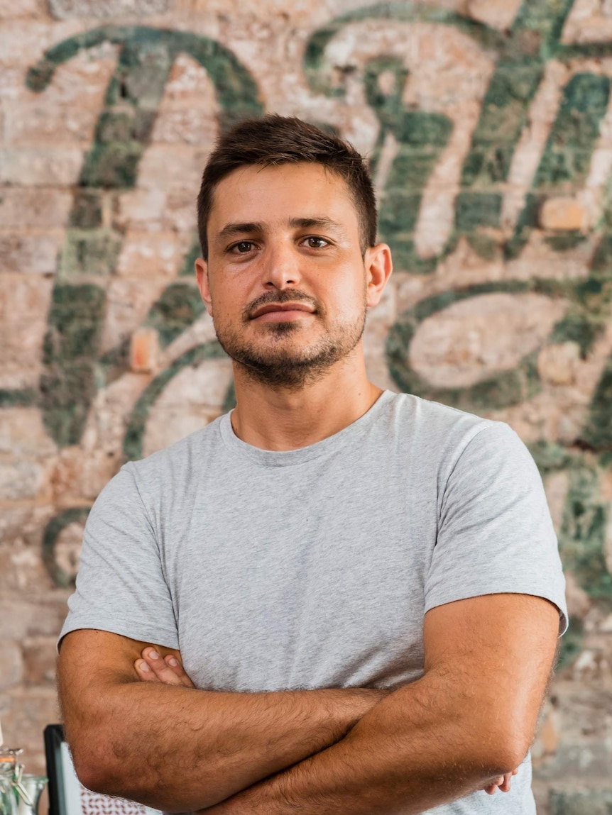 Cafe owner Dylan Peters in grey t-shirt stands with arms folded in front of a painted brick wall