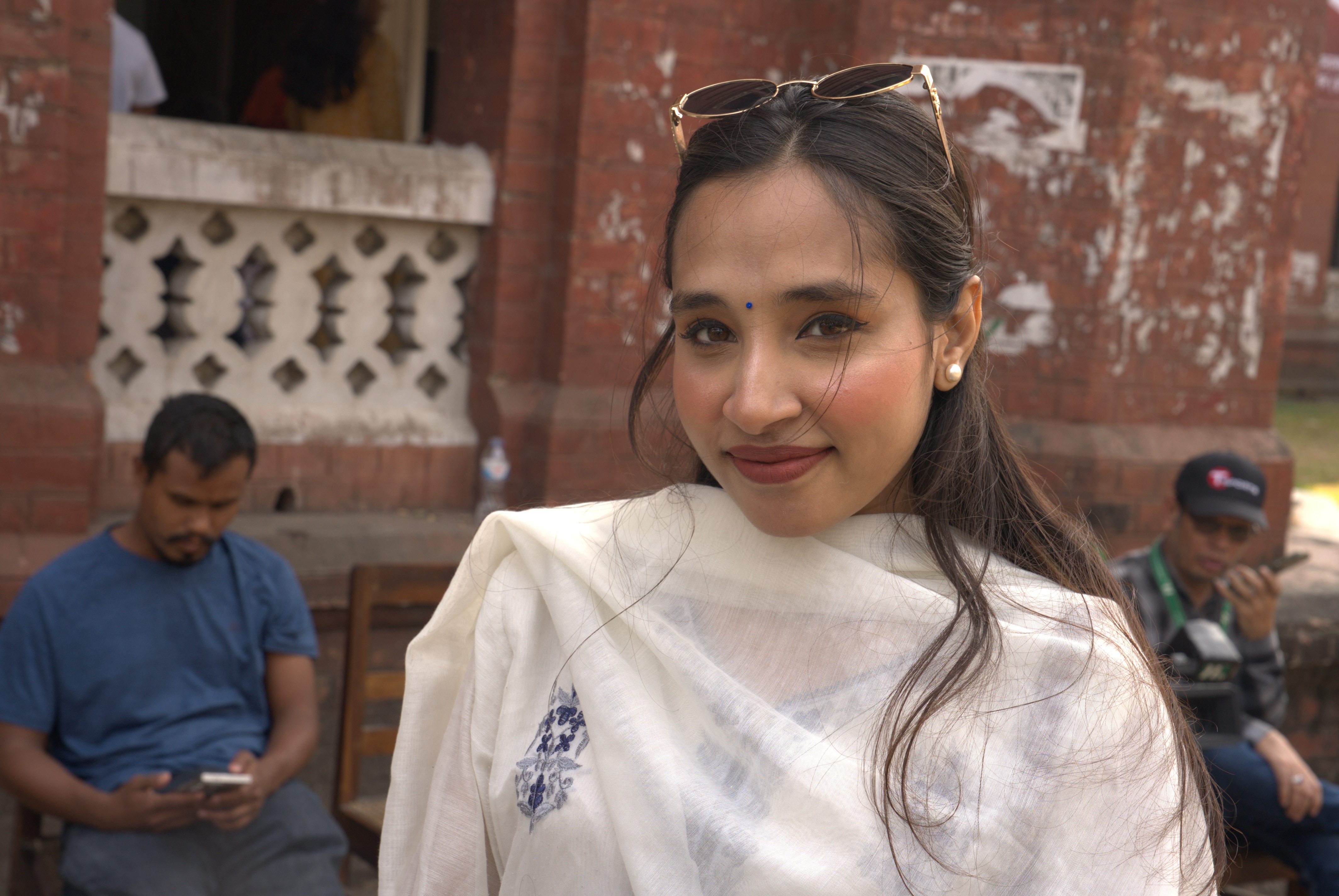 Una mujer sonríe a la cámara y usa gafas de sol en la cabeza mientras está parada frente a una pared roja.