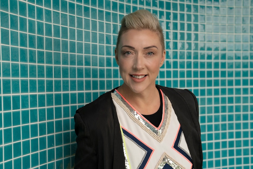 A woman with short blonde hair stands in front of an turquoise tiled wall.