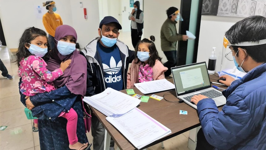 A family wearing a mask seeing a camera at a desk with a health worker.