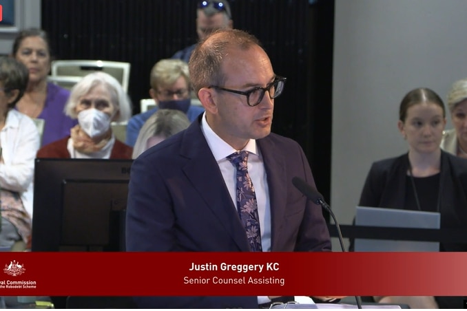 Greggery behind a desk and microphone, in front of seated onlookers, speaks