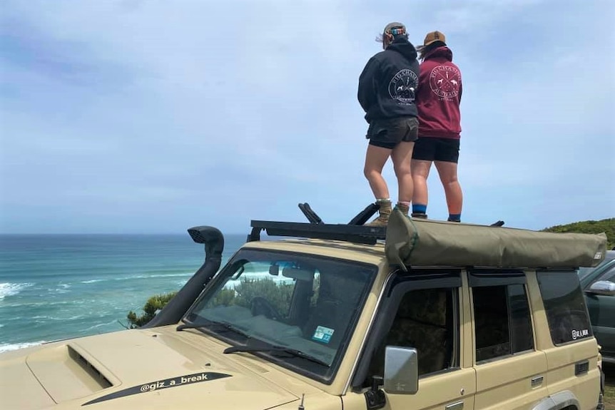 Two young women pictured from behind standing on the roof of a car and looking at the ocean.