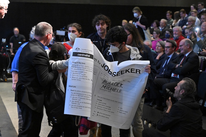 Masked protesters holding a JobSeeker banner interrupt a packed lecture hall.
