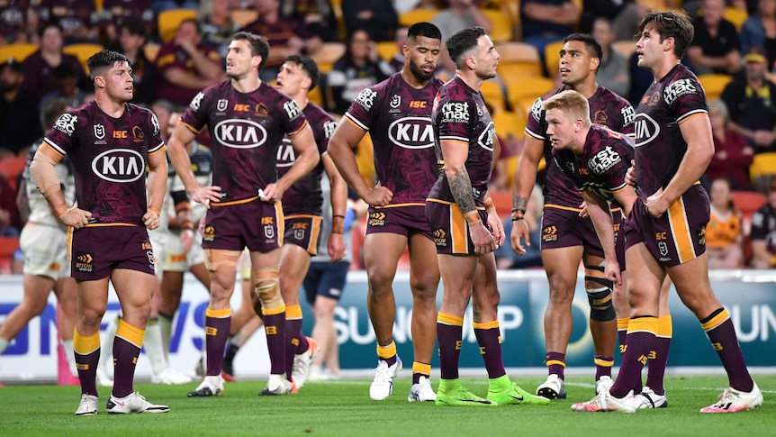 A group of NRL teammates stand looking dejected after a try by their opposing team in a game.