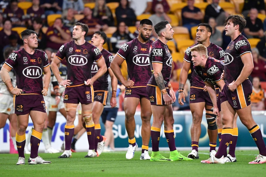 A group of NRL teammates stand looking dejected after a try by their opposing team in a game.