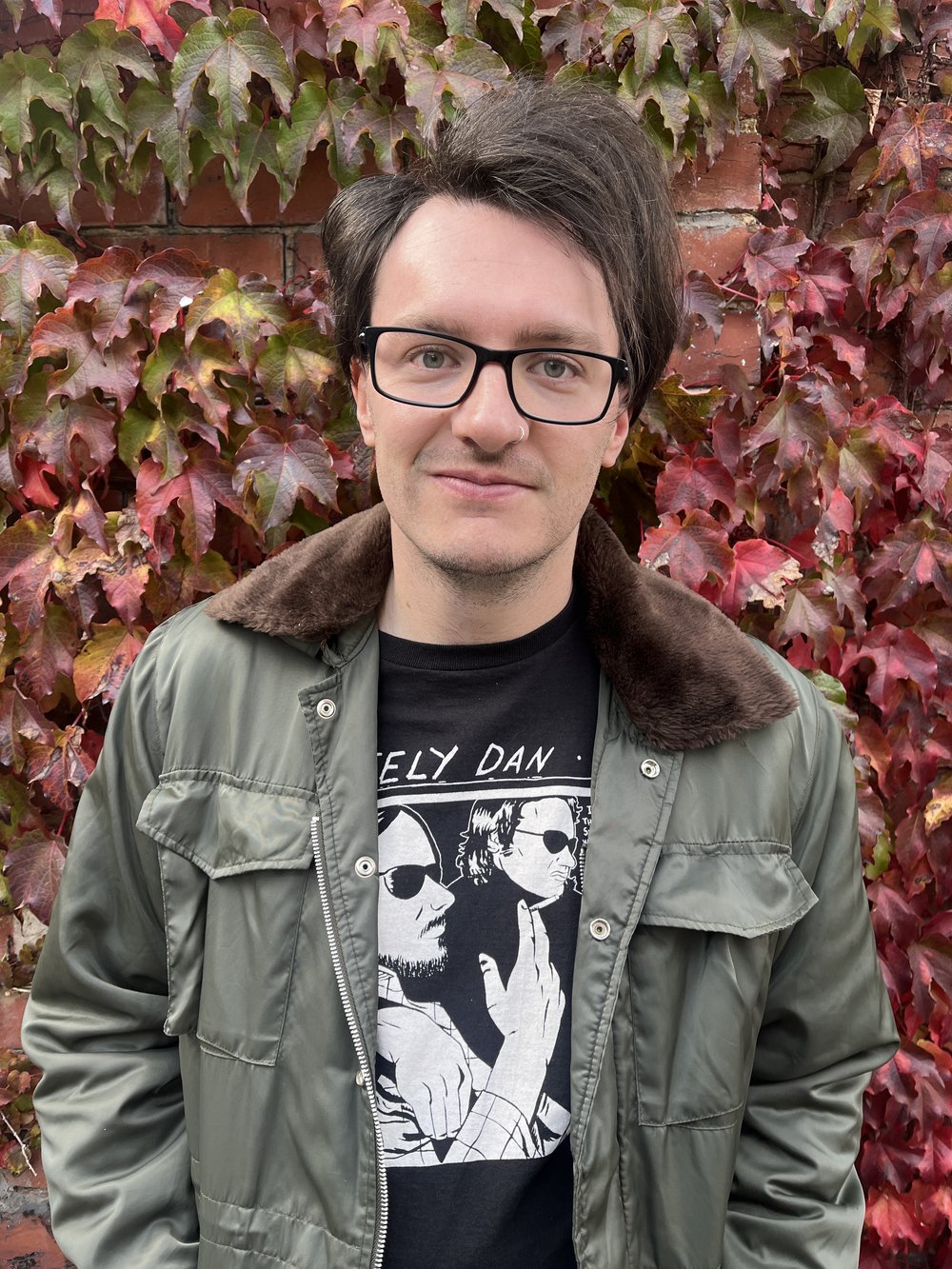 A man with brown hair and black rimmed glasses stands in front of a red and green leaf covered brick wall.