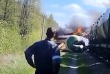 A young man in blue overalls stands with his hands on his hips as he watches a derailed train carriage burn.