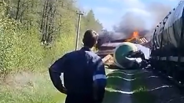 A young man in blue overalls stands with his hands on his hips as he watches a derailed train carriage burn.