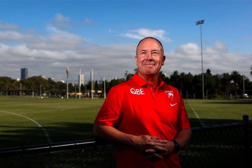 A man in a Sydney Swans polo smiles and looks at the sky in front of Sydney city