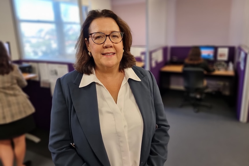 A woman stands in an office wearing glasses, a white collared shirt and grey jacket.