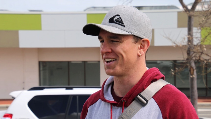 A man wearing a cap and hoodie and carrying a bag stands in front of a car smiling.