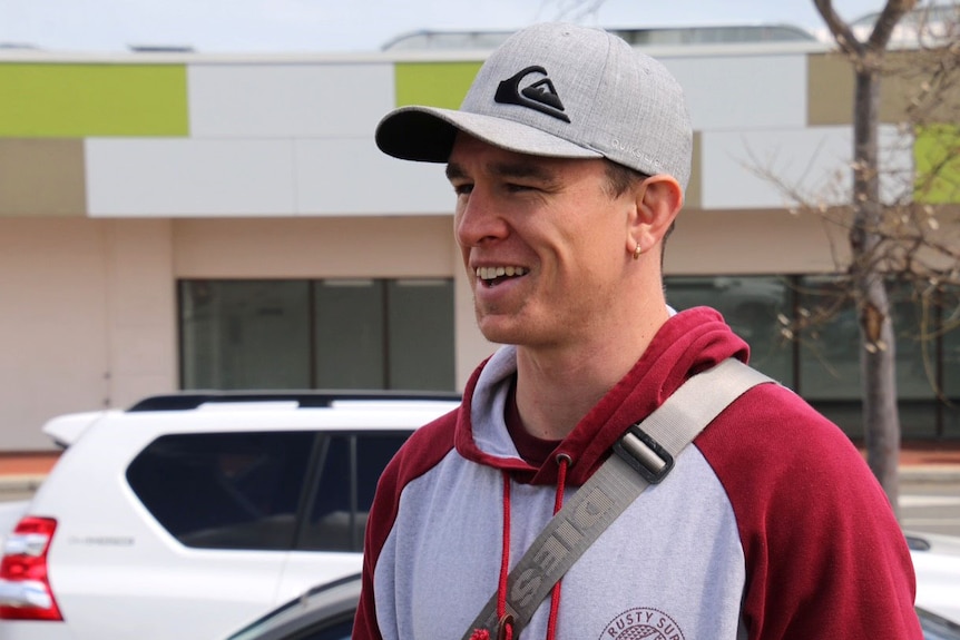 A man wearing a cap and hoodie and carrying a bag stands in front of a car smiling.