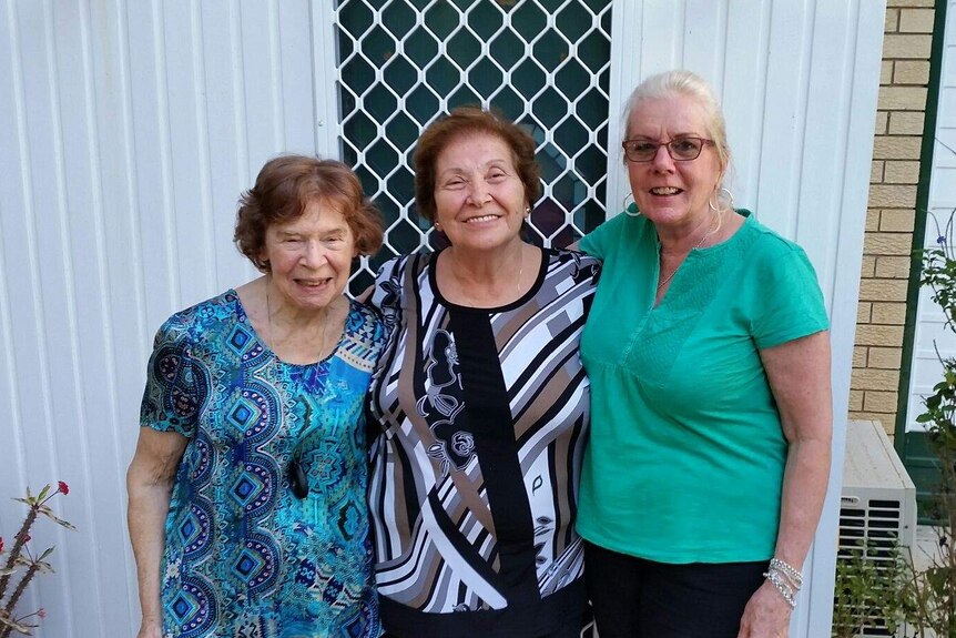 A woman wearing a personal alarm pendant stands with two other women in front of a sliding screen door.