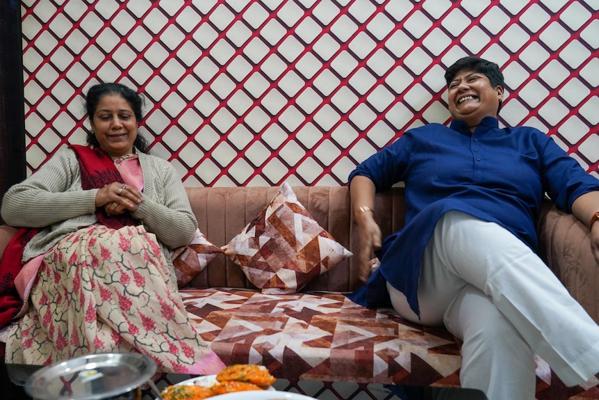 An older woman in a sari and a woman with close cropped hair sit together laughing on a couch