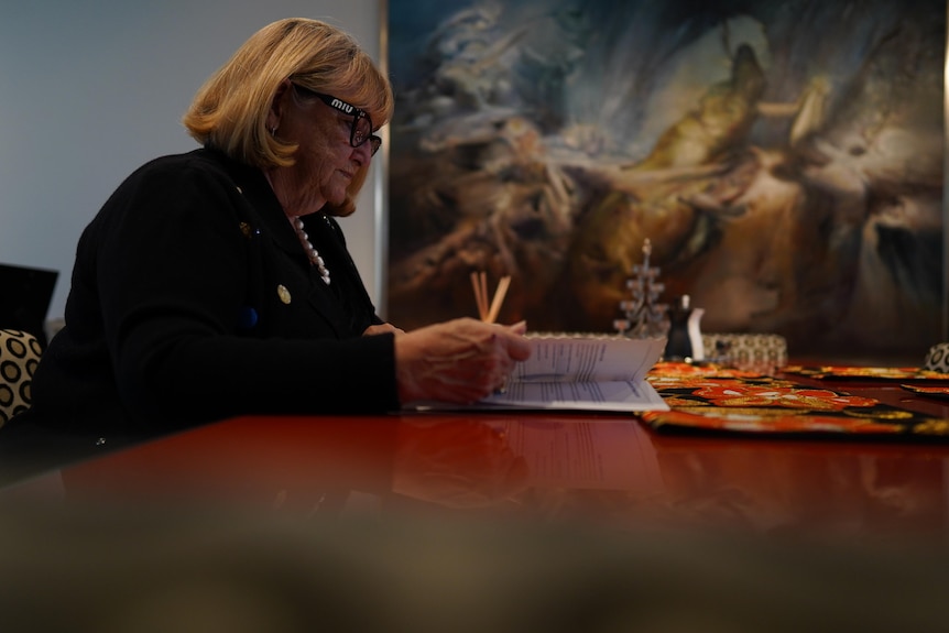 A woman sits at a desk in front of a painting.
