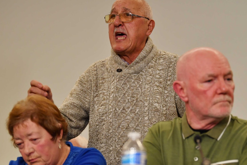 An emotional older man stands and speaks as two people sit in front of him