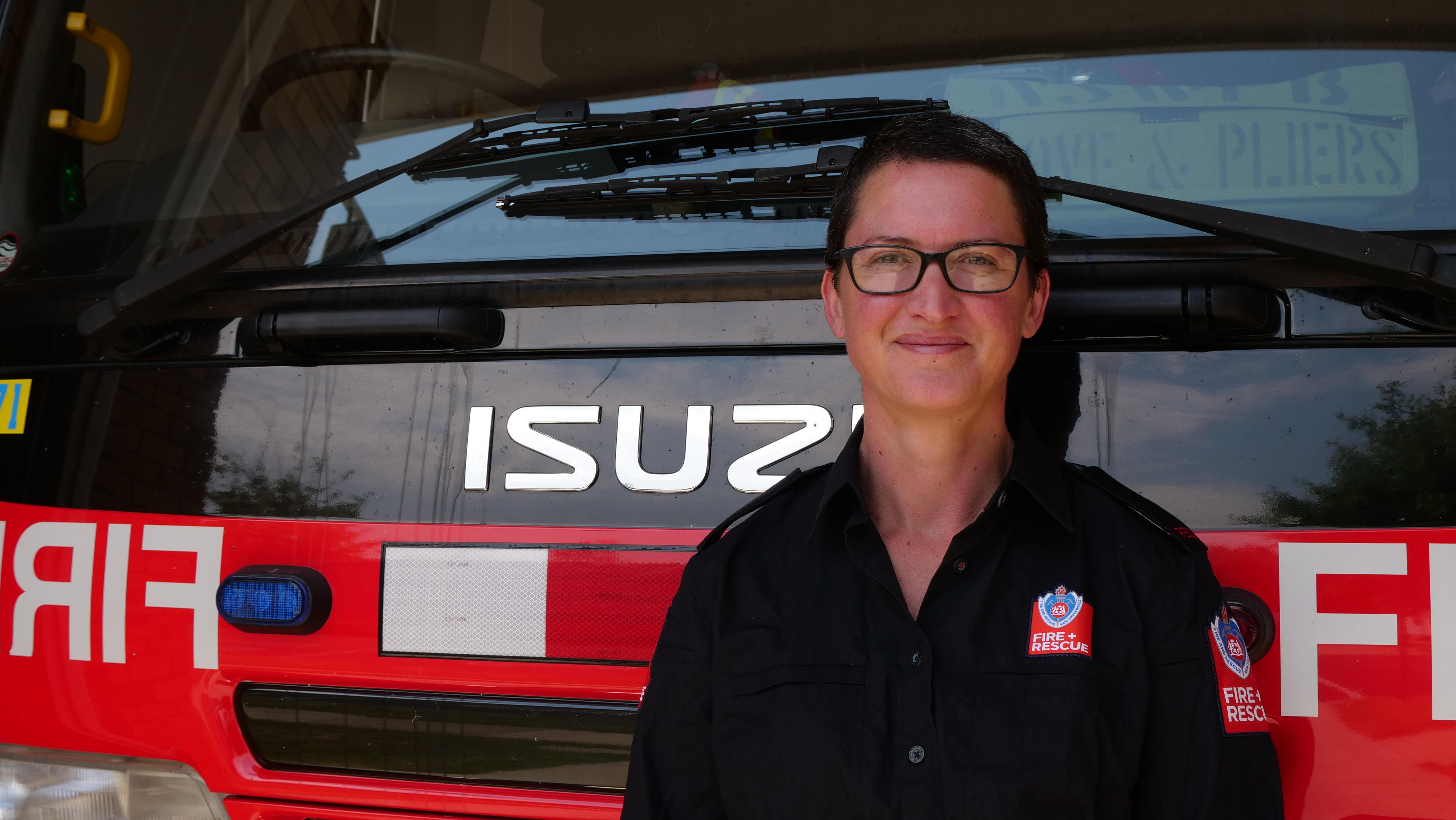 A female firefighter in a black uniform stands in front of a red fire truck. 